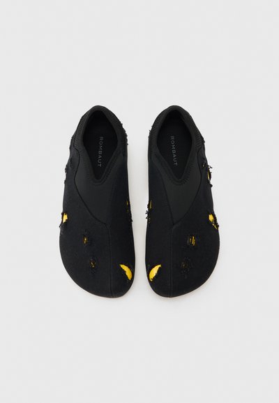 Ζευγάρι μαύρα παπούτσια slip-on με σκόπιμες κίτρινες σχισμές στο ύφασμα και μαύρο ελαστικό περιθώριο, φωτογραφημένα από πάνω πάνω σε γκρι φόντο.