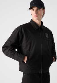 Jachetă neagră cu fermoar frontal, material texturat, guler pliat și buzunare laterale. Prezintă un patch cu logo pătrat pe piept.