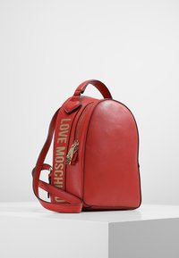 Červený kožený batoh s zlatým zipem, horní rukojetí, nastavitelnými popruhy a nápisem "LOVE MOSCHINO" vytištěným na bočním popruhu zlatými písmeny.