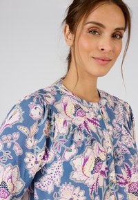 Blusa floral de color azul claro con patrones botánicos en púrpura y rosa, cierre de botones y detalles fruncidos en los hombros.