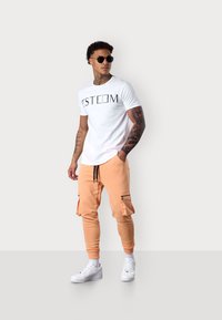 Weißes T-Shirt mit "ESTEEM"-Druck, kombiniert mit hellorangefarbenen Cargo-Jogginghosen mit Reißverschlusstaschen und weißen Sneakers. Schwarze Sonnenbrille inklusive.