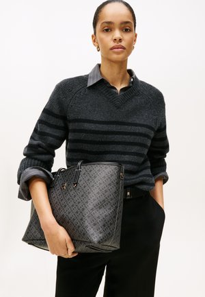 Grå mønstret tote taske med sorte detaljer, holdt af en person i en sort stribet sweater. Har en tekstureret overflade og en struktureret form.