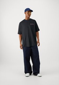 Sort oversized t-shirt med korte ærmer og rødt logo på brystet, parret med løse mørkeblå denimbukser og hvide sneakers.