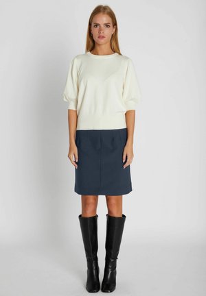 Žena stojaca, oblečená v bielom svetri s krátkym rukávom, tmavomodrej sukni po kolená a čiernych vysokých topánkach, na jednofarebnom svetlom pozadí.