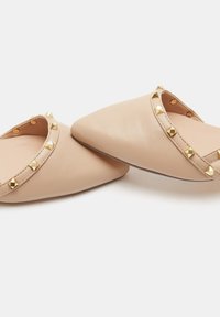 Zapatos sin cordones de puntas afiladas en tono marrón con un acabado de cuero suave y tachuelas en forma de pirámide en tono dorado a lo largo del borde superior. Diseño de suela plana.