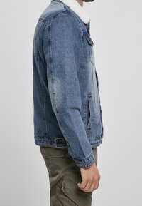 Hombre con chaqueta de denim azul y cuello de sherpa blanco, y pantalones cargo verdes oliva, de pie de perfil contra un fondo plano.