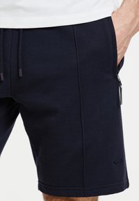 Pantalones cortos azul marino hechos de una tela suave, con cintura ajustable con cordón, bolsillo lateral con cremallera y detalle de logo bordado cerca del dobladillo.