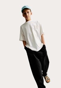 Vit t-shirt med kort ärm och texturerad detalj på framsidan, ihopparad med lösa svarta byxor och beige sandaler. Avslappnad keps i ljusblå.