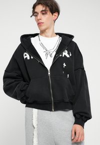Sweatshirt preto com zíper, de silhueta oversized, com frente desgastada, texto gráfico branco e bolsos espaçosos. Usado por cima de uma t-shirt branca.