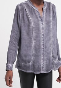 Blusa grigia a maniche lunghe con collo alto, tasche laterali e cuciture a contrasto. Realizzata in tessuto leggero e testurizzato.