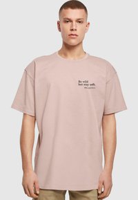 Lys rosa oversized bomull t-skjorte med korte ermer, med svart tekst, "Vær vill, men forbli myk. #lykke," på forsiden.