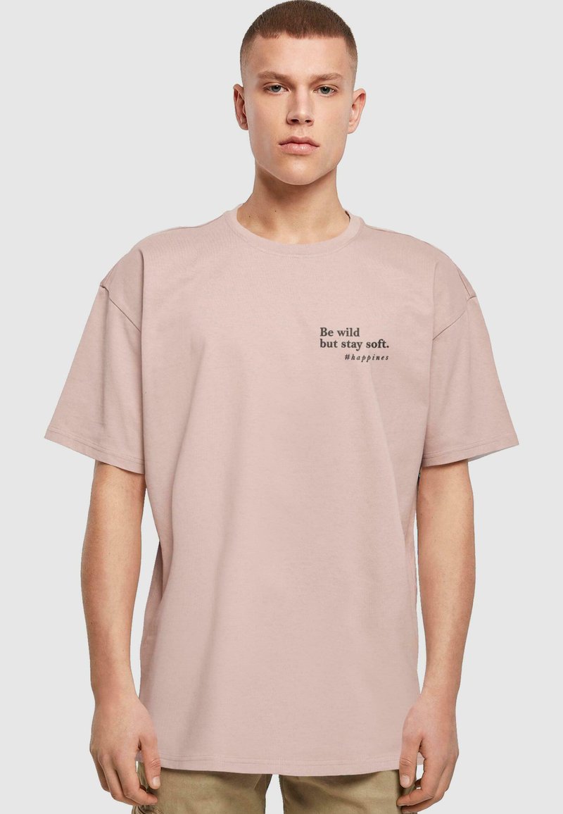 Lys rosa oversized bomull t-skjorte med korte ermer, med svart tekst, "Vær vill, men forbli myk. #lykke," på forsiden.
