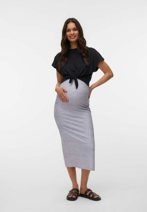 Gravid kvinde med langt mørkt hår iført sort bundet top, grå tætsiddende midi nederdel og sorte sandaler, smilende med hænderne på maven og hoften.