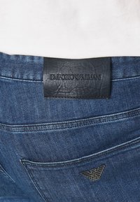 Džíny z denimu s tmavě modrým líhnutím, kožená etiketa s ražbou "EMPORIO ARMANI" a malým kovovým logem orla na zadní kapse.