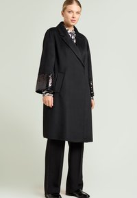 Svart lång ullkappa med vida ärmar, dekorerad med paljettaccent, snedskuren krage och framfickor, kombinerad med svarta byxor och skor.