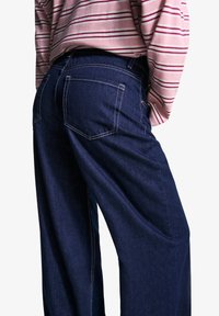 Temno modri jeans širokih nogavic z vidnimi zadnjimi žepi, belim šivanjem in sproščenim krojem, v kombinaciji s pajčolanasto rožnatim vrhom.