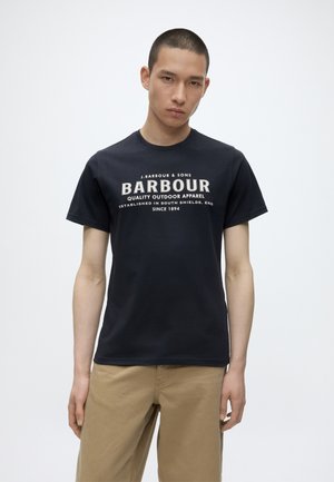 Νεαρός άνδρας που φοράει μαύρο T-shirt με το λογότυπο Barbour και μπεζ παντελόνι, στέκεται μπροστά σε απλό λευκό φόντο.