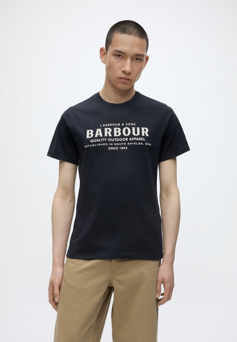 Νεαρός άνδρας που φοράει μαύρο T-shirt με το λογότυπο Barbour και μπεζ παντελόνι, στέκεται μπροστά σε απλό λευκό φόντο.