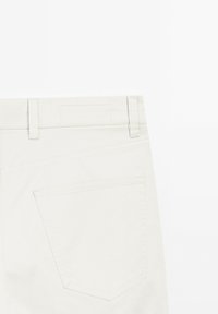 Beige bomullbyxor med låg midja, bälteshällor och en enda bakficka med en enkel rektangulär form och sömnadsdetaljer.