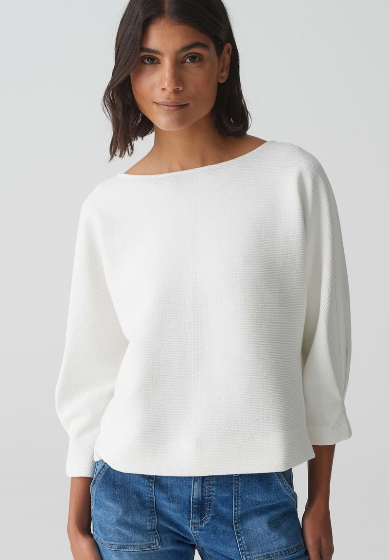 Pull blanc texturé avec un large col bateau et des manches amples, associé à un jean en denim bleu.