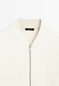 Veste bomber blanche zippée avec un col montant. Présente une fermeture à glissière argentée et des coutures soignées. L'étiquette indique "Massimo Dutti, fabriqué en Turquie."