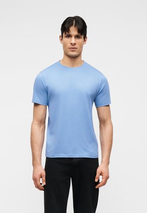 Άνδρας με κοντά σκούρα μαλλιά, φοράει ανοιχτόγαλανό κοντομάνικο μπλουζάκι και μαύρο παντελόνι, στέκεται μπροστά σε ένα απλό ανοιχτόχρωμο φόντο.