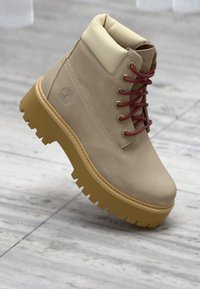 Μπεζ δερμάτινο μποτάκι αστραγάλου με χοντρή ταμπά σόλα, επενδεδυμένο κολάρο και κόκκινα κορδόνια, εμφανίζεται σε παστέλ γκρι πλακάκια.