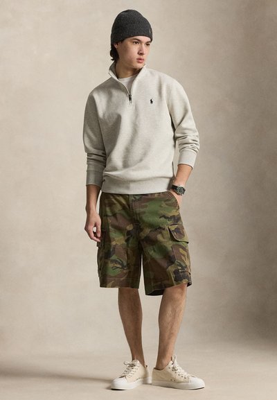 Jonge man draagt een grijze quarter-zip trui, een donkere gebreide muts, camouflage cargoshorts, witte sneakers en een polshorloge, staand met één hand in de zak.