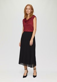 Haut bordeaux à col drapé associé à une jupe plissée noire avec un ourlet transparent. Le modèle porte des escarpins noirs à bout pointu avec des brides autour de la cheville.