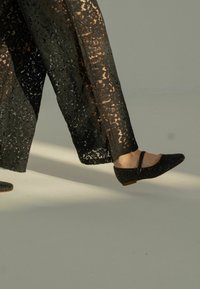 Zapatos planos negros con punta afilada y tira, hechos de un material texturizado con purpurina, llevados con pantalones de encaje negro translúcido.