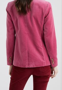 Růžový sametový blazer s upraveným střihem, klopovým límcem a zlatými knoflíky na rukávech. Hladká textura s těsným střihem.
