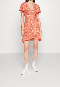 Červené květinové zavinovací šaty s krátkými volánkovými rukávy, výstřihem do V a rozšířeným lemem, doplněné bílými kotníkovými teniskami a ponožkami.