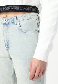 Calças de ganga de denim azul claro com uma cintura alta, apresentando um cós preto rotulado "LAGERFELD JEANS". O tecido exibe uma textura desbotada.