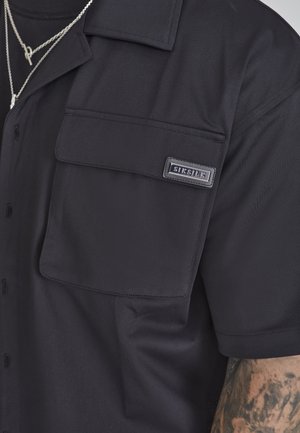 Schwarzes kurzärmeliges Hemd mit Klappentasche auf der Brust, beschriftet mit "SIKSILK", getragen mit silbernen Kettenhalsketten und sichtbarem tätowiertem Arm.