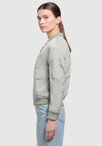 Jachetă bomber verde deschis cu mansete și guler ribelate, dotată cu buzunare cu fermoar și panouri laterale; textură netedă și design ajustat.