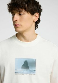 Ljusbeige sweatshirt med en rektangulär grafisk tryck av en klippkust i dimmiga blå och grå nyanser. Slät bomullstyg, rundad hals.