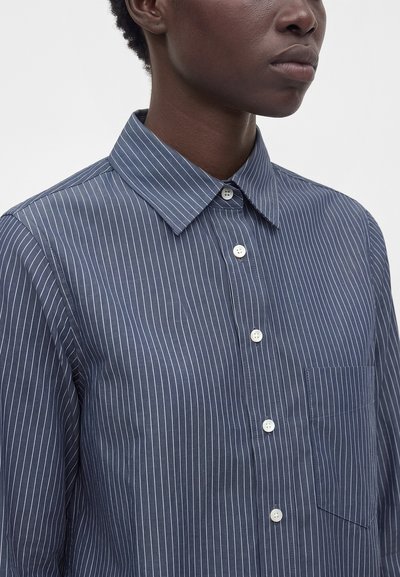 Personne portant une chemise bleu marine à fines rayures, boutonnée, avec un col pointu et une poche poitrine, montrée du cou au milieu du torse.