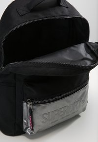 Musta kangaskantoreppu, jossa on harmaa vetoketjullinen etutasku. Ominaisuuksiin kuuluu sileä rakenne, logoyksityiskohta ja sisäinen vetoketjulla varustettu osasto.