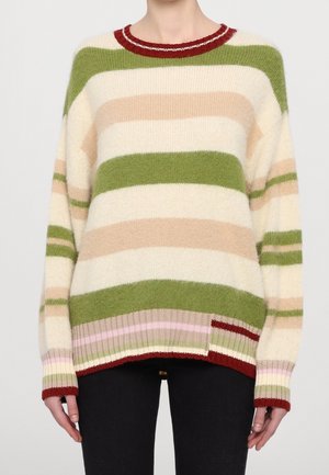 Personne portant un pull en maille ample à manches longues avec des rayures horizontales vertes, beiges, crème, roses et rouges, associé à un pantalon noir.