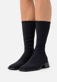 Botas negras de tobillo hechas de un tejido elástico, con punta afilada, tacón bloque y un diseño elegante que se extiende por encima del tobillo.