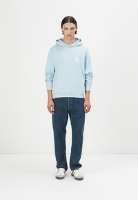 Lys blå hettegenser med kengurulomme, laget av bomull, med ribbede mansjetter og hem. Kombinert med mørkeblå jeans og hvite joggesko.