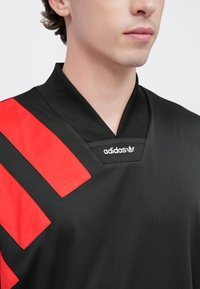 Čierny atletický dres s červenými diagonálnymi pruhmi, výstrihom do V a bielym logom adidas na golieri. Hladká, syntetická textúra.