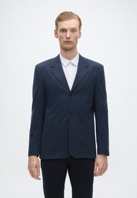 Blazer azul-marinho feito de tecido suave, apresentando um único bolso, dois bolsos frontais e dois botões pretos. Usado sobre uma camisa branca.