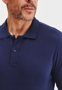 Granatowa koszulka polo z teksturowanej tkaniny, z dwoma guzikami przy kołnierzu i wytłoczonym logo po lewej stronie.