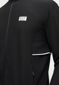Μαύρο μπουφάν με φερμουάρ και λευκό λογότυπο EA7 Emporio Armani στο στήθος και λευκή λεπτομέρεια περιγράμματος στο καπάκι της πλαϊνής τσέπης.