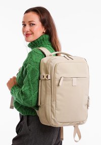 Bežový batoh s hladkým povrchem, více zipy a nastavitelnými popruhy, nošený přes zelený pletený svetr.
