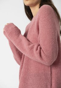 Pulover tricotat roz, cu o textură moale și pufoasă, având mâneci lungi și mansete elasticizate. Design minimalist, cu o croială lejeră.