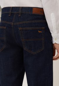 Temno modri jeans s oranžnim šivom, z usnjenim pasom in majhnim logotipom psa na zadnjem žepu.