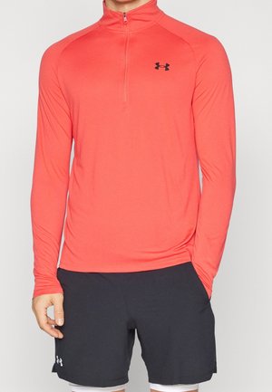 T-shirt de sport rouge à manches longues avec un design à demi-zip et un logo noir sur la poitrine, associé à un short noir et un sous-vêtement blanc.