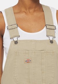 Salopettes kaki avec bretelles réglables, clips en métal et une poche avant avec un patch logo Dickies. Tissu en coton avec une texture lisse.